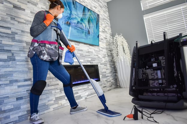 Cleaner using a steam mop to clean under furniture that's been moved to expose hidden dirt.